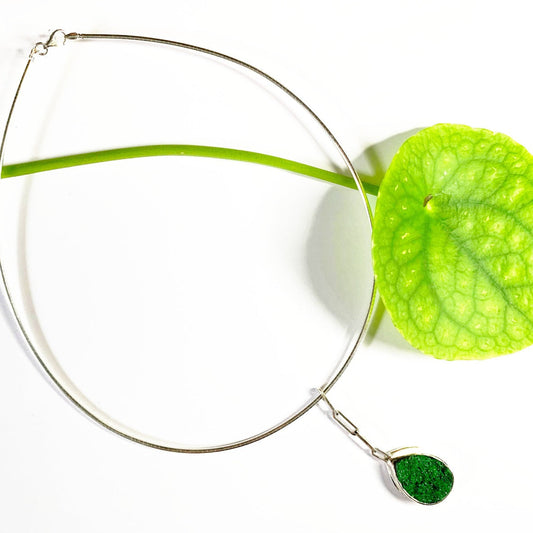 Et si vous accrochiez un fragment de roche à votre cou ? Ce collier en argent de 40 cm présente au centre une uvarovite druzy sertie clos pour la protéger des chocs. Sa forme druzy fait qu'elle est recouverte de minuscules inclusions brillants au soleil. Ce pendentif est long de 5 cm, accroché par des chaînons rectangles en argent. Vue de face.